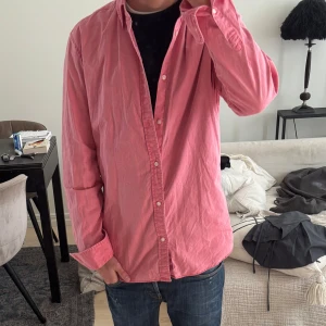 Rosa skjorta  - Säljer en klassisk rosa skjorta med knäppning framtill och krage. Skjortan har lång ärm och en avslappnad passform, perfekt att styla med jeans eller chinos. Enkel och stilren design utan synliga mönster eller loggor.