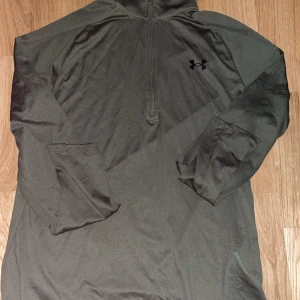 Olivgrön träningströja med dragkedja från Under Armour - Säljer en olivgrön långärmad träningströja från Under Armour med halv dragkedja framtill. Perfekt för träning eller utomhusaktiviteter. Tröjan har en sportig look och är tillverkad i ett syntetmaterial som andas.