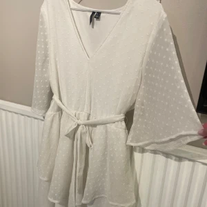 Vit omlottklänning med volang från Vero Moda - Superfin vit klänning i storlek L med prickigt mönster, v-ringning och volangdetaljer. Klänningen har trekvartsärm och knyte. Perfekt till sommaren eller studenten🤍