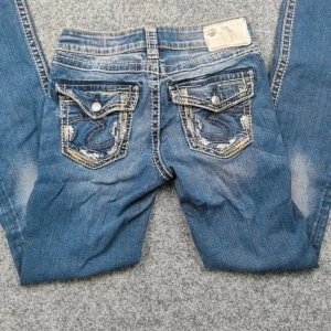 Blå jeansbyxor med broderade bakfickor från Miss Me - Säljer ett par blå jeansbyxor från  fluid denim med snygga broderade detaljer och knappar på bakfickorna. Jeansen har en klassisk femficksdesign och slitningar för en cool look. Perfekta för dig som gillar detaljer och unik stil. Köpa på vinted men var för små! Lånade bilder p alla förutom sista, de är slim bootcut