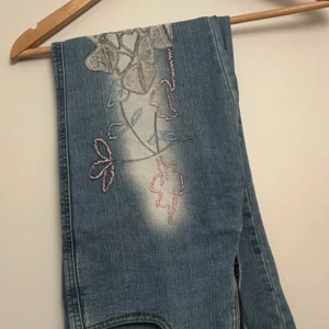 Blå broderade jeansbyxor från Wanili - Jättesöta, somriga, blå jeansbyxor med broderade blommor och blad i rosa och vit på framsidan. Byxorna är bootcut och har ljusa slitningar längs benen.