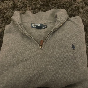 Grå halvzip tröja från Polo Ralph Lauren - Säljer en stilren grå halvzip-tröja från Polo Ralph Lauren med klassisk blå logga broderad på bröstet. Tröjan har lång ärm, ribbade muddar och är tillverkad i mjuk bomull. Perfekt för lager-på-lager och enkel att matcha.