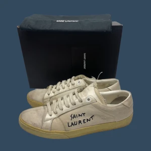 Saint Laurent skor - Snygga beige sneakers från Saint Laurent med handskriven logga på sidan. Skorna har en klassisk design med snörning och en robust gummisula. Perfekta för en stilren och avslappnad look. Skick 8 av 10! 🙌