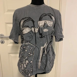 Grå t-shirt med glittrigt tryck från Zara - Säljer en grå t-shirt från Zara med ett stort tryck av två personer med glittriga detaljer och pärlor på glasögonen. T-shirten har korta ärmar och en avslappnad passform. Perfekt för dig som gillar unika motiv och lite extra bling.