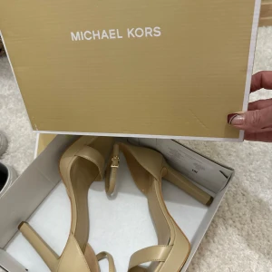 Beige pumps från Michael Kors - Säljer ett par stilrena beige pumps från Michael Kors med öppen tå och smal klack. Skorna har en elegant design med en tunn rem runt vristen och guldfärgad detalj. Perfekta för dig som vill ha en klassisk och snygg sko.