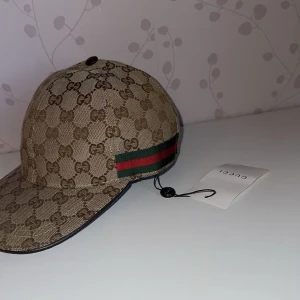 Beige keps från Gucci med monogram - Säljer en beige keps från Gucci med klassiskt GG-monogram och gröna/röda ränder på sidan. Kepsen har böjd skärm och justerbar rem baktill. Perfekt för dig som gillar lyxiga accessoarer och vill sticka ut.