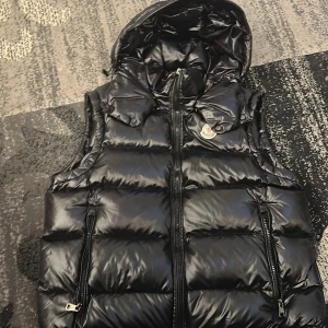 Svart dunväst från Moncler - Snygg svart dunväst från Moncler med glansig finish och huva. Västen har dragkedja framtill och två fickor med dragkedja. Klassisk Moncler-logga på bröstet och serietidningsdetalj på insidan.