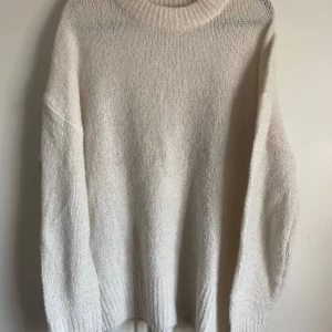 Vit stickad tröja med rund hals - Mysig vit stickad tröja med bred ribbad rund halsringning. Tröjan har lång ärm och en avslappnad passform, perfekt för kyliga dagar. Enkel och stilren design som passar till det mesta.