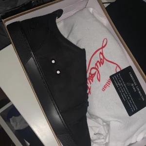 Svarta sneakers från Christian Louboutin - Säljer ett par helsvarta sneakers från Christian Louboutin med diskret logga broderad på hälen och klassisk röd sula. Skorna har snörning och är tillverkade i mocka med detaljer i skinn. Perfekta för dig som gillar stilrena och exklusiva sneakers.