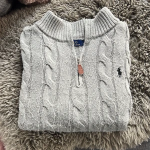 Polo Ralph Lauren half zip kabelstickad tröja - Säljer en ljusgrå half zip kabelstickad tröja från Polo Ralph Lauren. Tröjan har lång ärm, ribbade muddar och den klassiska broderade loggan på bröstet. Tröjan är  noppig. Perfekt för dem som vill se lite finare ut i vardagen
