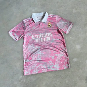 Real Madrid Special Edition Pink Dragon T-Shirt - Säljer en rosa Real Madrid fotbollströja från Adidas med vitt drakmönster och vita detaljer. Tröjan har korta ärmar, vit krage och klubbmärke på bröstet. Emirates Fly Better-logga på framsidan. Aldrig använd helt ny, passar inte mig därför säljer jag den