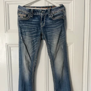 Lågmidjade bootcut jeans från Rock Revival - Säljer detta blå par bootcut jeans från Rock Revival med snygga broderade bakfickor och vita sömmar! Väldigt skönt material och perfekt för dig som söker några unika jeans🥰Stl 29, säljer då de är lite för stora..