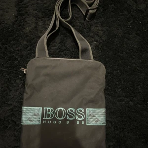 Svart axelväska från Hugo Boss - Snygg svart axelväska från Hugo Boss med justerbar rem och dragkedja upptill. Väskan har ett stort huvudfack och ett mindre innerfack med dragkedja. Perfekt för att bära med sig det viktigaste i vardagen.