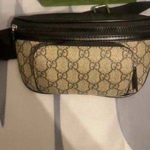 Beige magväska - Säljer en beige magväska med klassiskt GG-mönster och svarta detaljer. Väskan har dragkedja upptill och ett extra fack framtill. Perfekt för att bära det viktigaste med stil.