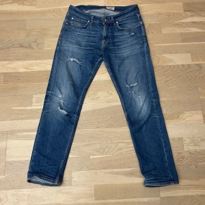 Blå jeans från Tiger of Sweden - Säljer ett par blå jeans från Tiger of Sweden med slitna detaljer och klassisk femficksdesign. Snygga slitningar och storleken är L30 W31