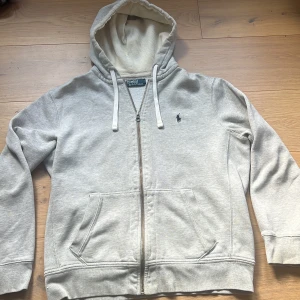 Ljusgrå hoodie från Polo Ralph Lauren - Säljer en ljusgrå hoodie från Polo Ralph Lauren med dragkedja och huva. Tröjan har två fickor framtill, justerbara snören i huvan och den klassiska broderade loggan på bröstet. Perfekt för en avslappnad stil.