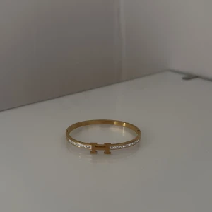 Guldfärgat armband med H-detalj - Snyggt guldfärgat armband med en H-formad detalj framtill och glittrande stenar längs ena sidan. Perfekt accessoar för att lyfta din outfit.