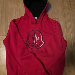 Röd hoodie från Moncler - Säljer en röd hoodie från Moncler med stor logga framtill. Tröjan har svart insida på huvan och klassisk passform. Perfekt för dig som gillar streetwear och vill ha något snyggt och bekvämt.