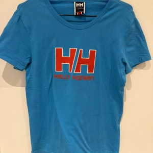 Blå t-shirt från Helly Hansen - Blå t-shirt från Helly Hansen med stort rött HH-tryck och logga framtill. Klassisk rund halsringning och korta ärmar. Perfekt för en sportig och avslappnad stil.