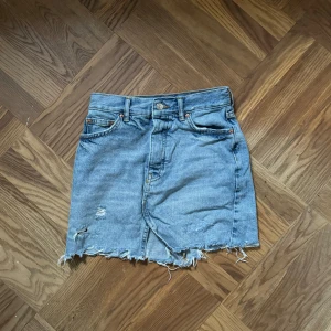 Blå jeanskjol  - Säljer en blå jeanskjol med rå, fransig nederkant och slitningar framtill. Kjolen har klassiska fem fickor och knappgylf. Storlek 34. Jag är ca 160 för referens. 🌸