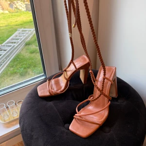 Bruna sandaler med snörning och klack - Säljer ett par trendiga bruna sandaler med fyrkantig tå, hög klack och långa flätade snören som kan lindas runt benet. Skorna har öppen tå och plattformsula framtill. Perfekta för att lyfta en somrig outfit.
