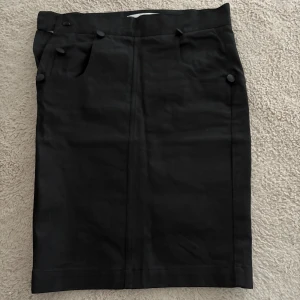 Svart pennkjol från Alexandra Fundin - Säljer en svart pennkjol från Alexandra Fundin med hög midja och dekorativa knappar framtill. Kjolen har fickor både fram och bak, och är i ett stilrent, klassiskt snitt. Perfekt för dig som gillar en enkel men snygg look.