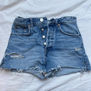 Blå jeansshorts med slitningar från ZARA - Säljer ett par blå jeansshorts från ZARA med hög midja och råa, slitna detaljer. Snyggt med en stor stickad tröja över midjan!! Shortsen har knappar framtill, klassiska fickor och fransig kant nertill. Perfekta för en avslappnad stil.