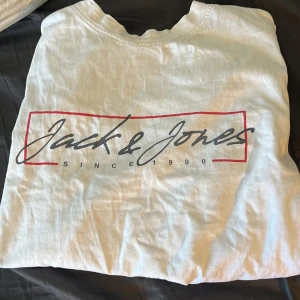 Vit t-shirt från Jack & Jones - Säljer en vit t-shirt från Jack & Jones med tryckt logga och texten 'Since 1990' på bröstet. Klassisk passform och rund halsringning med röd detalj på insidan av kragen. Perfekt till vardags.