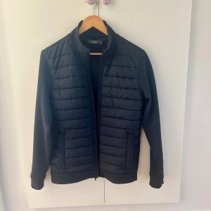 Svart quiltad jacka från Dressmann - Säljer en svart quiltad jacka från Dressmann med dragkedja framtill. Jackan har ståkrage, två fickor med dragkedja och släta ärmar. Perfekt för kyligare dagar och enkel att matcha med olika outfits.