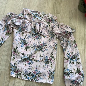 Blommig volangblus från Lindex - Säljer en ljusrosa blus från Lindex med blommigt mönster i blått, grönt och lila. Blusen har långa ärmar och volangdetaljer längs axlar och bröst. Perfekt för vår och sommar!
