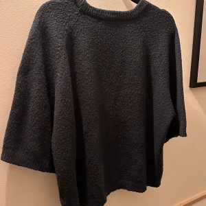 Mörkblå stickad ponchotröja - Mysig mörkblå stickad tröja med lös passform och trekvartsärmar. Perfekt att slänga på sig över en topp för extra värme. Enkel och stilren design utan detaljer.