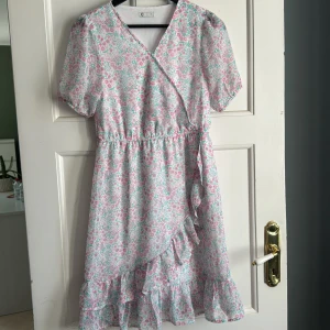 Blommig omlottklänning med volang - Söt kortklänning med blommigt mönster i ljusa pastellfärger. Klänningen har omlottmodell, korta ärmar och volangdetalj längs nederkanten. Perfekt för vår och sommar!