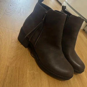 Bruna boots med dragkedja - Säljer ett par bruna boots med rund tå och dragkedja på sidan. Skorna har en platt sula och en liten klack, samt dekorativ rem vid dragkedjan. Perfekta för dig som vill ha en stilren och enkel look.