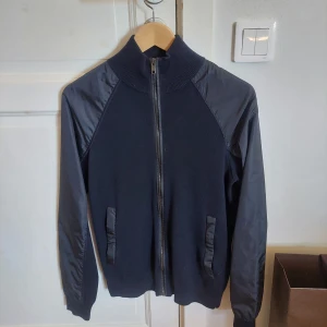 Prada cardigan - Marinblå prada cardigan i storlek 46. Den är gjord av 100% ylle, med polyamid applikationer på ärmarna. Det är en mycket klassisk design med en touch av tuffhet av ärmapplikationerna. Den är i nyskick förutom att tags o.s.v. saknas. Nypris 14500 kr.