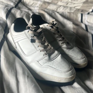 Vita sneakers med svarta detaljer - Snygga vita sneakers med svarta detaljer och beige snörning. Skorna har rund tå, platt sula och är tillverkade i syntetmaterial. Perfekta för en stilren och sportig look.