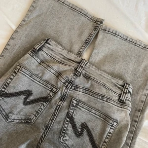 Grå jeans med svarta detaljer från Nelly - Säljer ett par grå jeans från Nelly med bootcut och svarta sicksack-detaljer på bakfickorna. Jeansen har hög midja, dubbla knappar och klassisk femficksdesign. Perfekta för dig som gillar en unik twist på dina jeans! Använd en gång, köpt för 699kr
