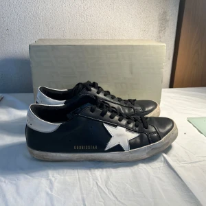 Svarta sneakers från Golden Goose - Säljer ett par svarta Golden Goose sneakers med vit stjärndetalj på sidan och vit häl. Skorna har snörning och en något smutsad platt sula för den klassiska GGDB-looken. Perfekta för dig som gillar streetstyle. Skicka 8/10