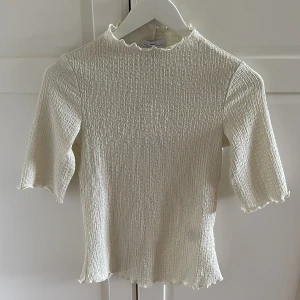 Vit ribbad topp från & Other Stories - Säljer en vit ribbad topp från & Other Stories med korta ärmar och hög hals. Toppen har en smockad struktur och vågiga kanter vid hals, ärmslut och nederkant. Perfekt att styla till många olika looks!