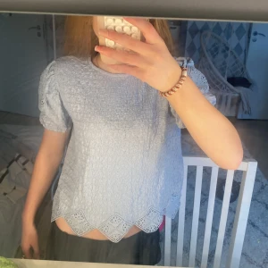 Ljusblå blus med spetsdetaljer från Zara - Säljer en ljusblå blus från Zara med korta puffärmar och spetsdetaljer vid ärmslut och nederkant. Blusen har en vågig struktur och en unik kant nertill med spets. 
