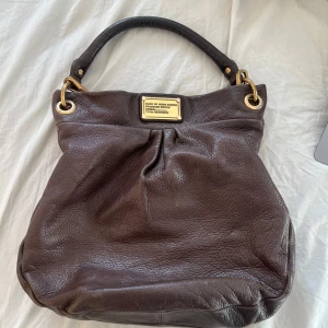 Mörkbrun handväska från Marc by Marc Jacobs - Säljer en mörkbrun handväska i skinn från Marc by Marc Jacobs med guldfärgade metalldetaljer och justerbart handtag. Väskan har en snygg logotypplatta framtill och ett innerfoder med svartvitt textmönster.