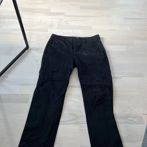 Svarta kostymbyxor från KappAhl - Klassiska svarta kostymbyxor från KappAhl med raka ben och diskreta bakfickor med knappdetalj. Byxorna har hällor för bälte och stängs med dragkedja och knapp framtill. Perfekta för en stilren look.