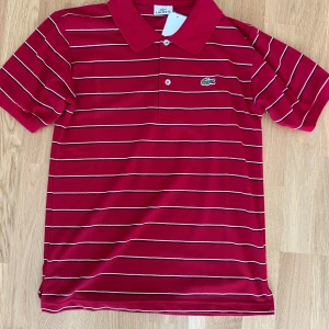 Röd randig pikétröja från Lacoste - Säljer en klassisk röd pikétröja från Lacoste med vita och mörkblå ränder. Tröjan har korta ärmar, krage och knäppning framtill samt den ikoniska krokodilloggan på bröstet.