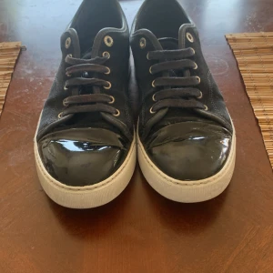 Svarta sneakers med lackad tå - Säljer ett par svarta sneakers från Lanvin med snygg lackad tå och vit sula. Skorna har snörning och är gjorda i mocka med detaljer i skinn. Perfekta för dig som vill ha en stilren men ändå unik look.