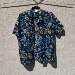 Blommig kortärmad skjorta från W. Shiner - Snygg kortärmad skjorta från W. Shiner med blå, turkosa och gula blommor på mörkblå botten. Skjortan har klassisk krage och knäppning framtill. Storlek M.