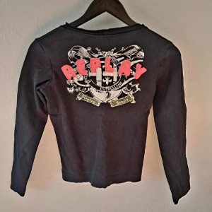 Svart långärmad tröja Replay - Svart långärmad tröja från Replay med v-ringning. På bröstet finns en liten broderad Replay-logga och på ryggen ett stort tryck med röda bokstäver och coolt emblem. Använd men bra skick, se bilder. Står L på lappen men passar storlek S. 