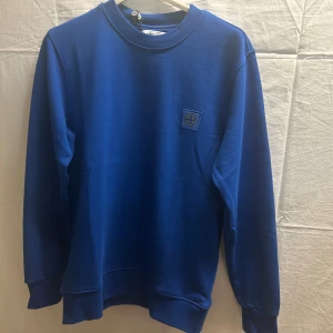 Blå sweatshirt från Stone Island - Snygg blå sweatshirt från Stone Island med klassisk logopatch på bröstet. Tröjan har rund hals, långa ärmar och ribbade muddar. Tillverkad i mjuk bomull som känns skön mot huden. Perfekt för dig som gillar stilrena och sportiga plagg.             Obs! Alla storlekar finns. Skriv önskad storlek efter köp