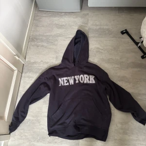 Svart hoodie med New York-tryck - Cool svart hoodie med stor huva och trycket 'NEW YORK' i vitt på bröstet. Klassisk känguruficka framtill och långa ärmar. Perfekt för en avslappnad streetstyle-look.