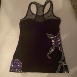 Metal Mulisha svart linnetopp mesh - Svart linnetopp från Metal Mulisha med meshparti upptill och brottarrygg. Tryck med lila rosor, dödskalle och smyckesdetaljer samt logga på ryggen. Snygg kontrast mellan det svarta tyget och de lila detaljerna. Perfekt för dig som gillar alternativ stil.