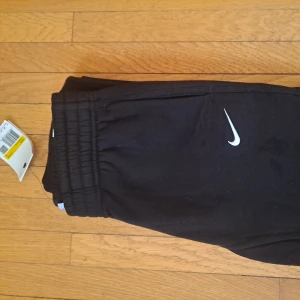 Svarta mjukisbyxor från Nike - Svarta mjukisbyxor från Nike med klassisk vit logga på ena benet. Byxorna har resår i midjan och raka ben, perfekta för chill eller träning. Mjukt material som känns skönt mot huden. Helt nya. Säljer de för att dem för för små. 