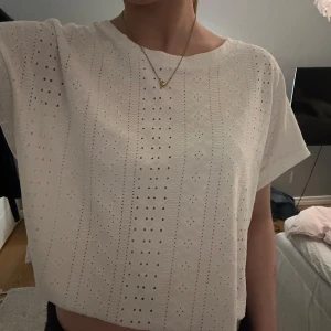 Vit croppad blus med broderade hål - Säljer en söt vit croppad blus med korta ärmar och broderade hål i ett diskret mönster. Perfekt för varma dagar och enkel att matcha med både jeans och kjol.